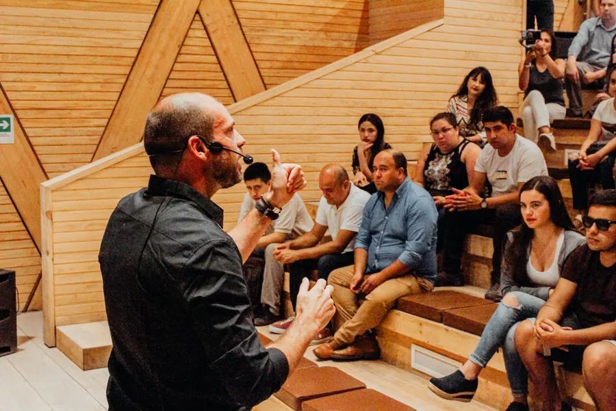 Felipe Bozzo, con micrófono de cintillo, conversa de cerca con un grupo de personas sentadas en las gradas de un anfiteatro de madera